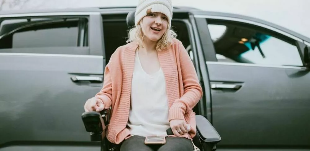 En dame sitter i rullestol utenfor en bil. Foto